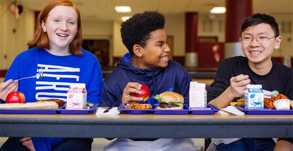 Three students sit at a metal cafeteria table eating hamburgers with cartons of milk on school lunch trays. credit: Healthy School Meals for All NY Kids